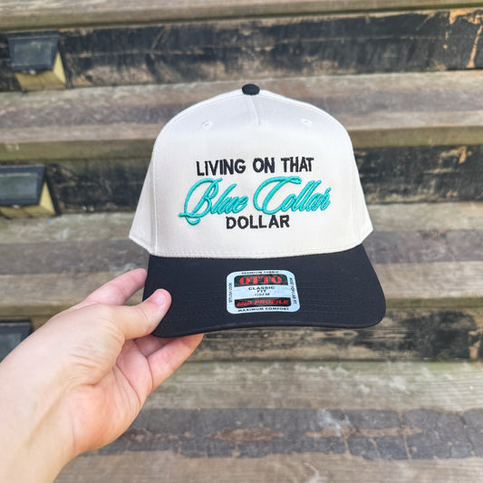 Cap with 'Living on that Blue Collar Dollar' text held by a hand against a wooden background