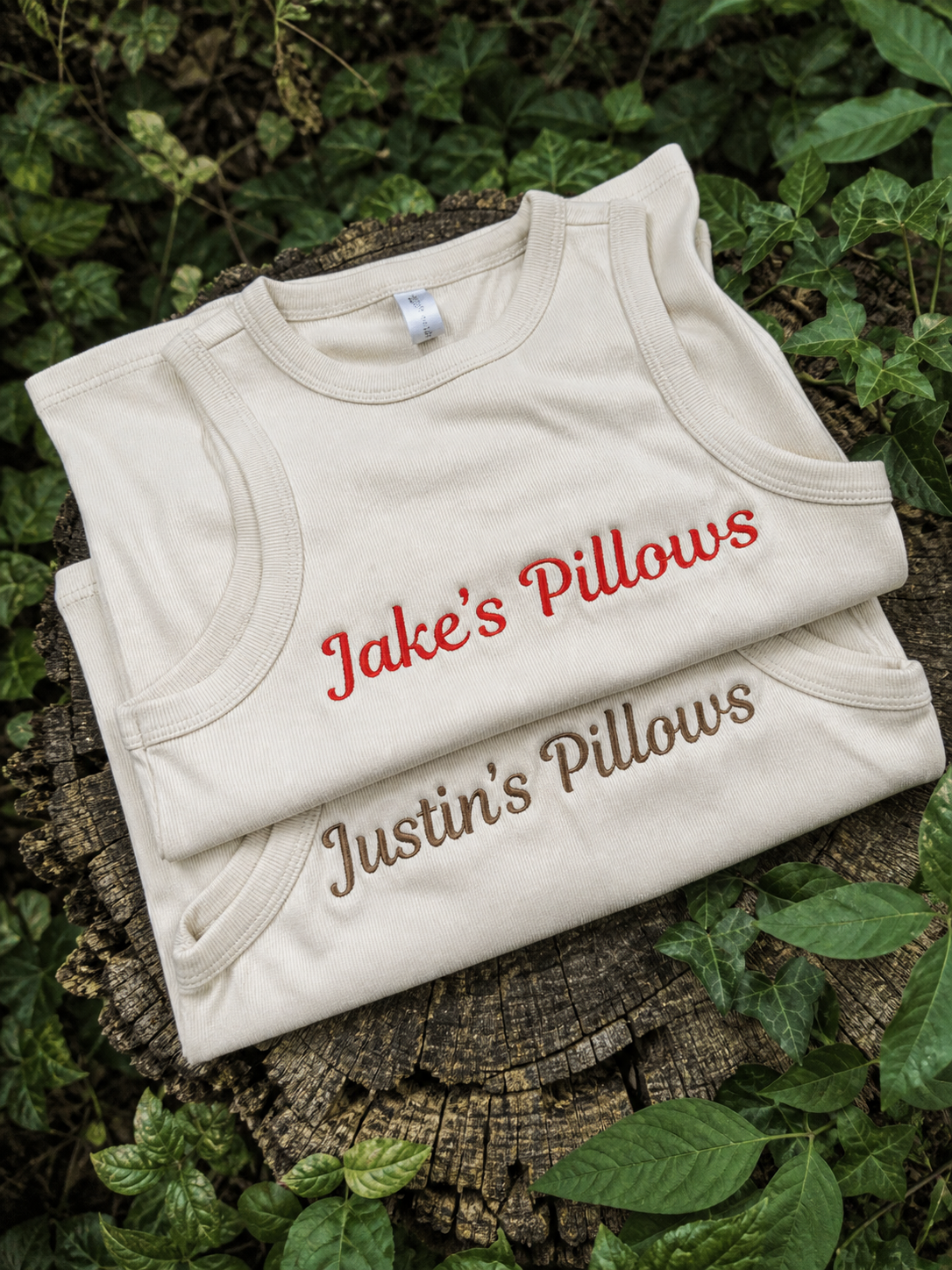 Folded beige tanks with embroidered names on a wooden surface surrounded by green leaves.