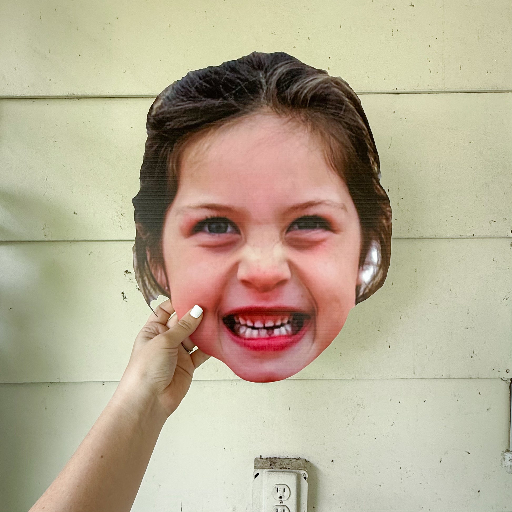 Cardboard cutout of a woman's face held up against a light-colored wall.