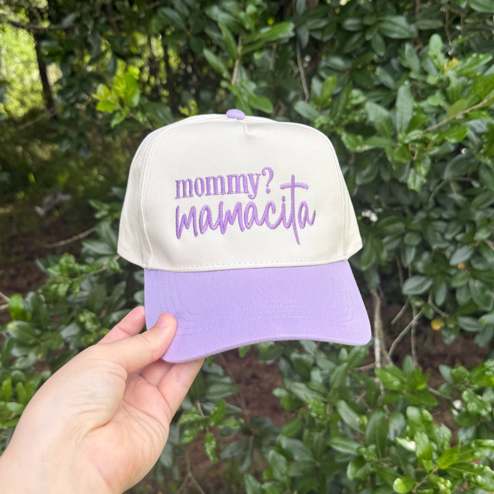 Cap with 'mommy? mamacita' text held in front of green foliage