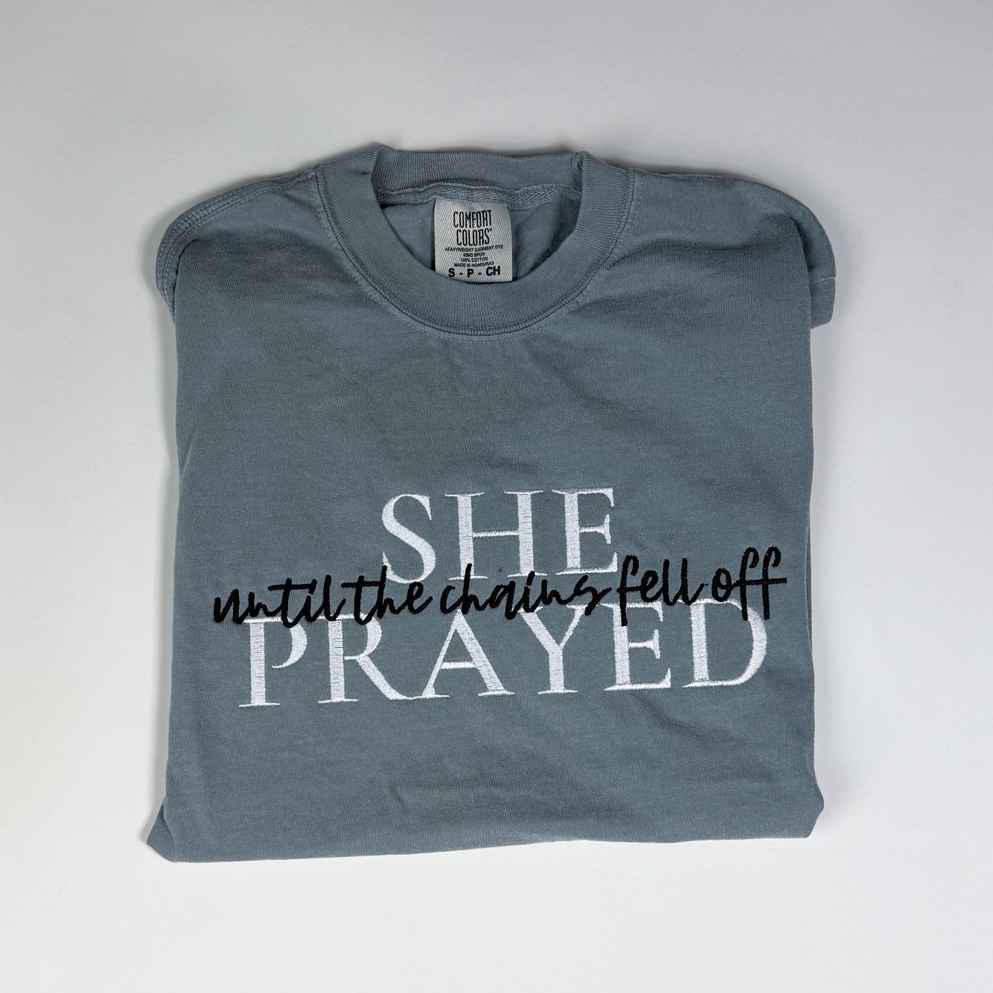 Folded grey t-shirt with text 'SHE PRAYED until the chains fell off' on a white background