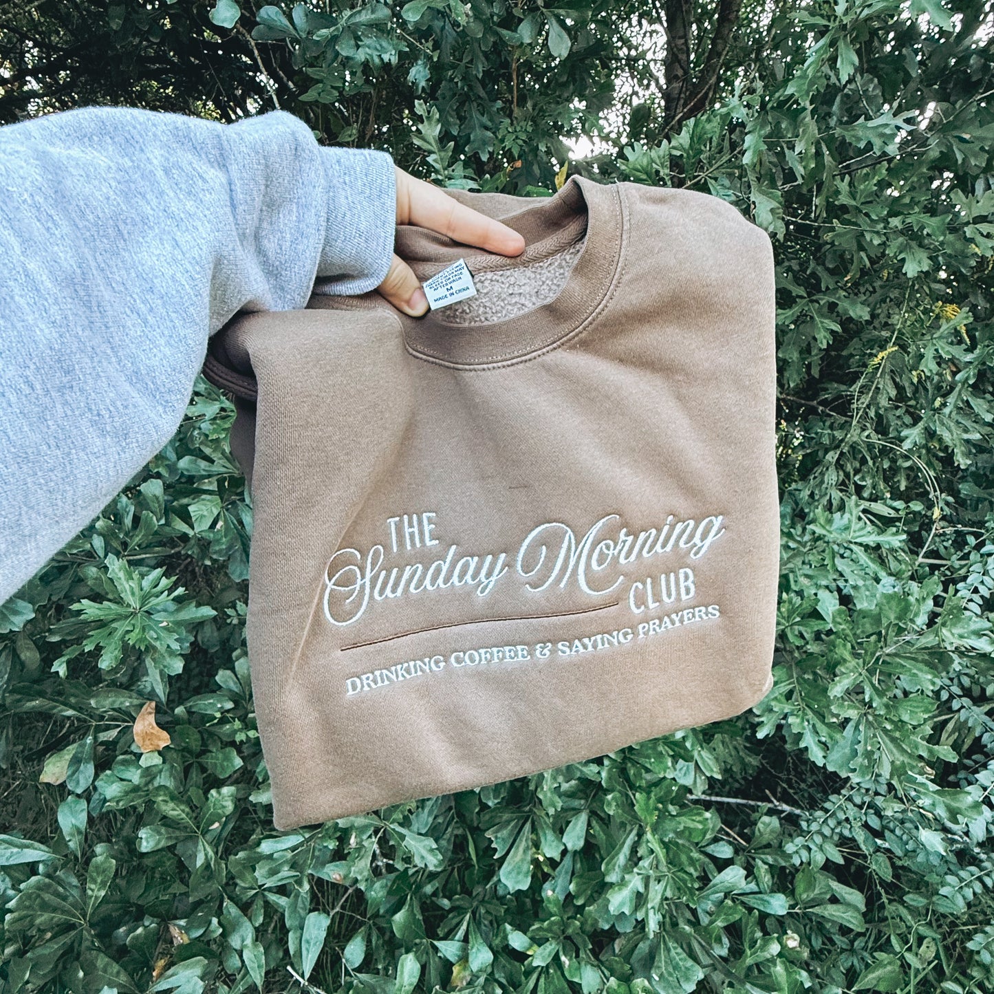 Beige sweatshirt with 'The Sunday Morning Club' text held against a green leafy background