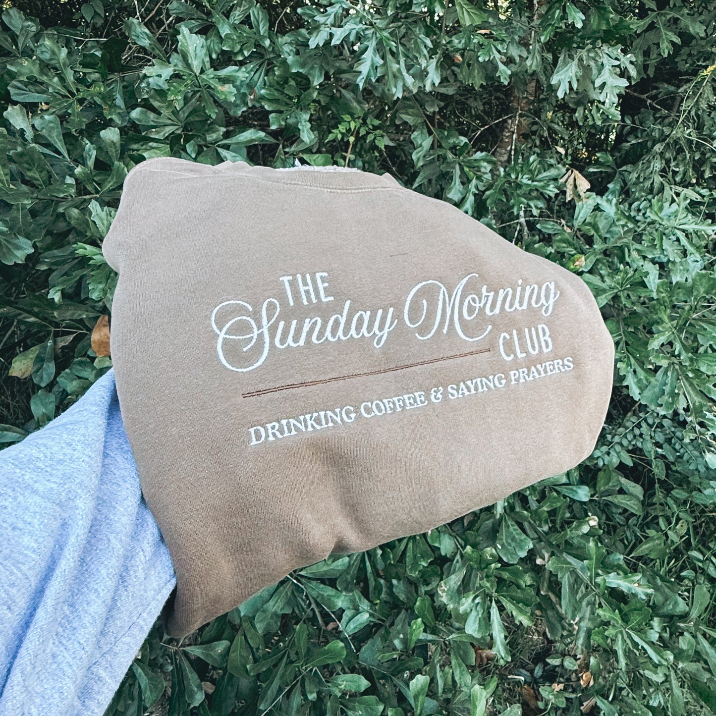 Brown sunday morning club sweatshirt being held in front of greenery