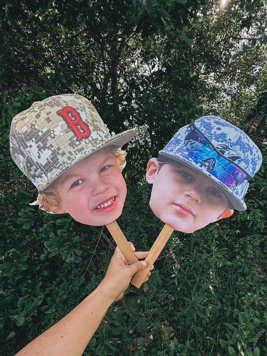Two childrens cutout face masks with printed faces against a green outdoor background